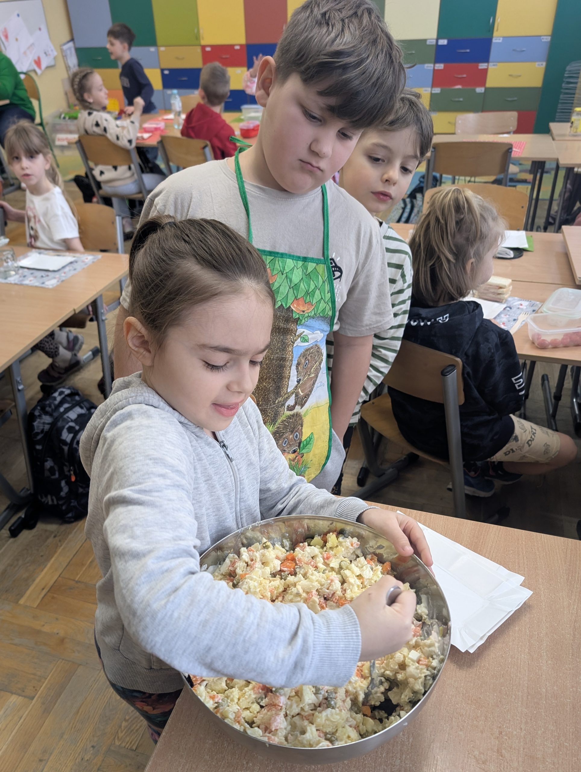 Dziewczynka miesza dużą miskę sałatki jarzynowej, obok niej stoją dwaj chłopcy przyglądający się z zainteresowaniem. W tle widać klasę pełną dzieci oraz kolorowe szafki.