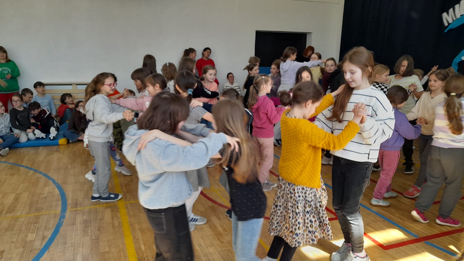 Grupa dzieci tańczy w parach na sali gimnastycznej trzymając się za ręce. W tle siedzą dzieci obserwujący taniec.