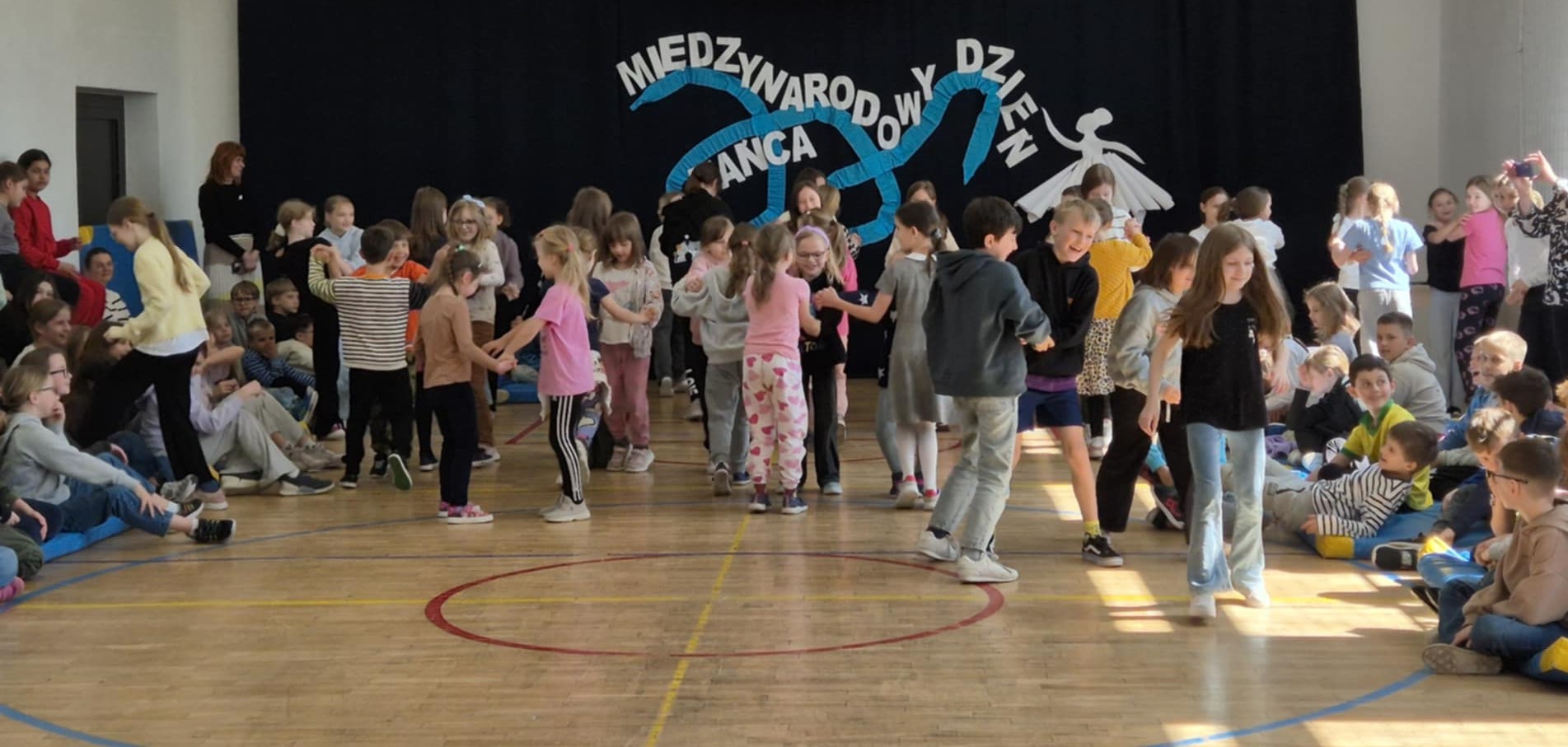 Grupa dzieci tańczących w parach w sali gimnastycznej. W tle inne dzieci siedzące na materacach oraz napis na scianie "Międzynarodowy Dzień Tańca"