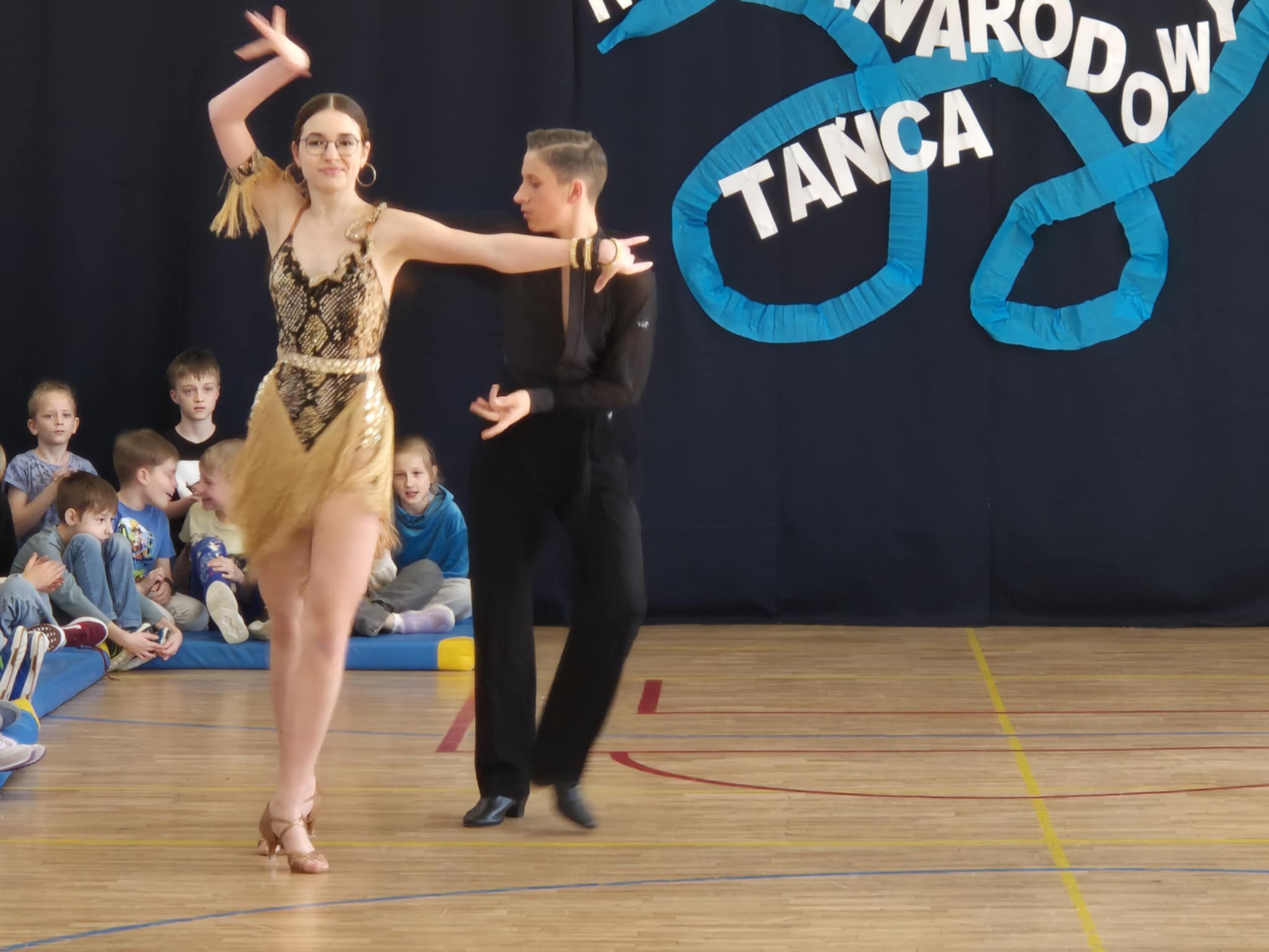 Para tancerzy podczas występu na sali gimnastycznej. W tle siedzi grupa dzieci obserwujących występ. 