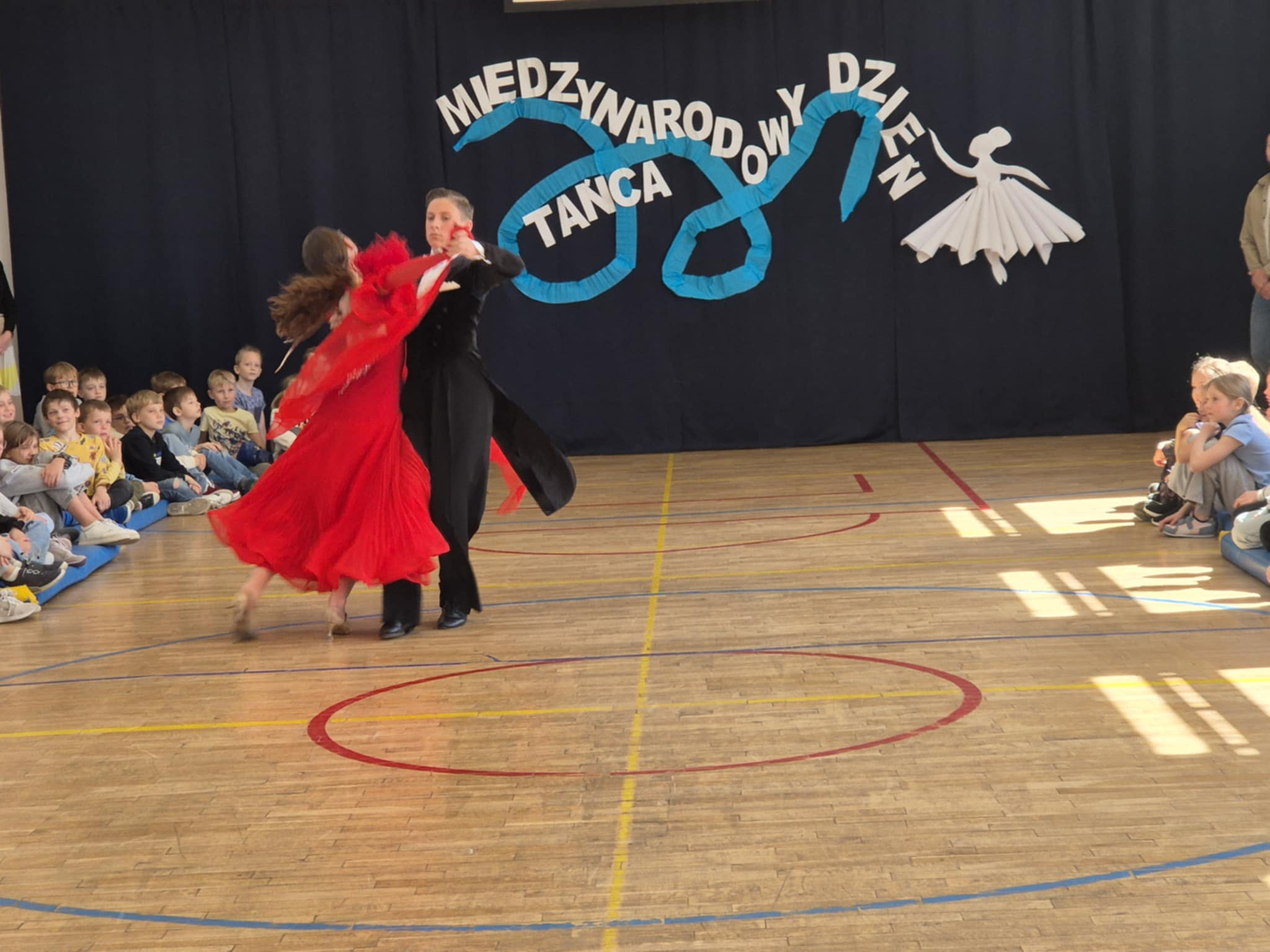 Para tancerzy podczas występu na sali gimnastycznej. W tle siedzi grupa dzieci obserwujących występ. 