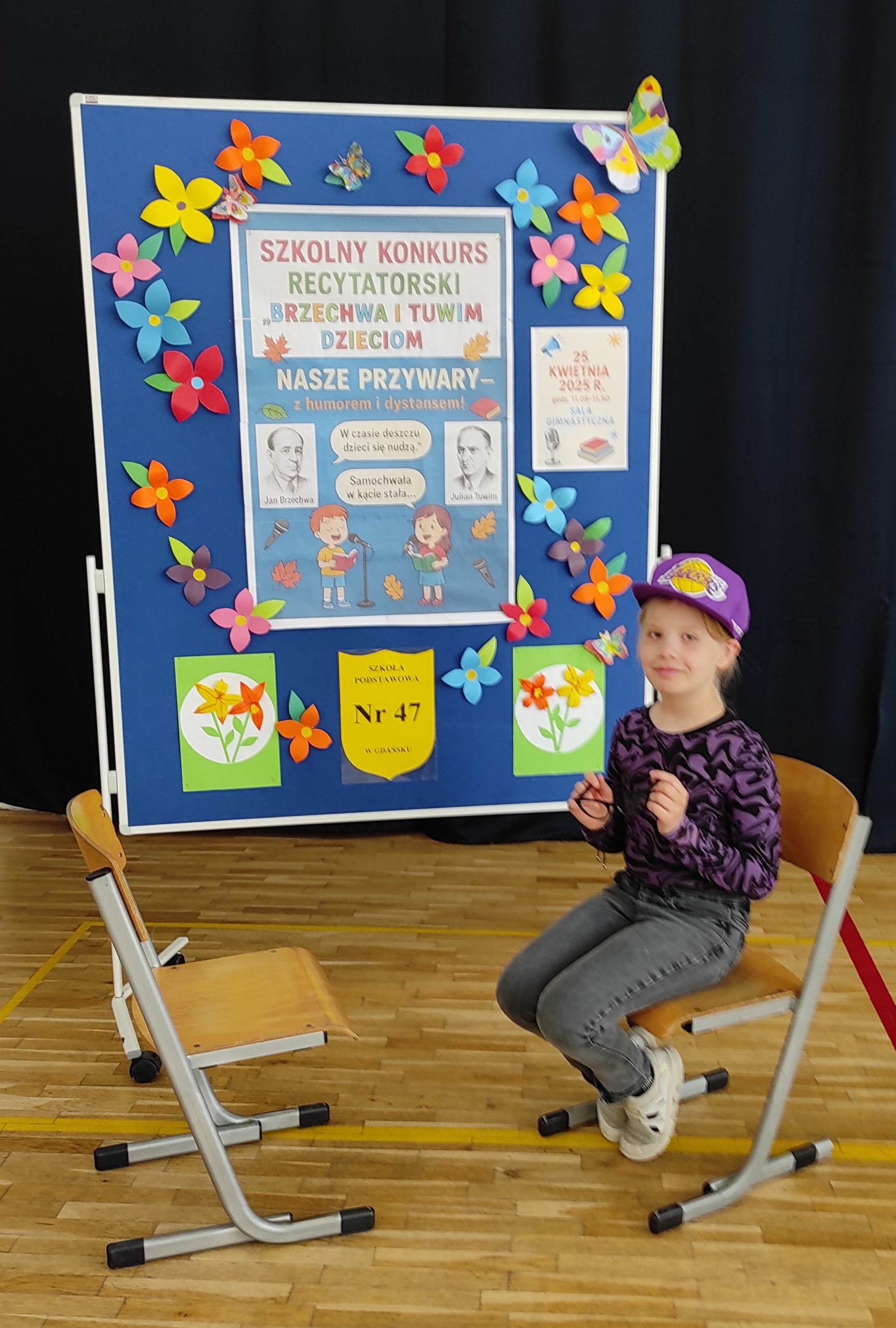 W tle tablica informacyjna z napisem: KONKURS RECYTATORSKI "Brzechwa i Tuwim dzieciom" - NASZE PRZYWARY i kolorowymi kwiatami. Na pierwszym planie dwa krzesła ustawione przodem do siebie. Na jednym z nich siedzi dziewczynka w czapce z daszkiem, w dłoniach trzyma okulary.