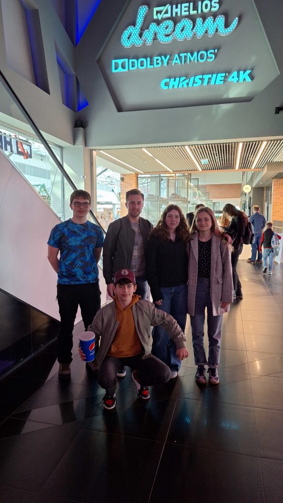 Na zdjęciu jest 5 osób w lobby kina, w tym dwie dziewczyny, dwóch chłopaków i mężczyzna. Stoją pod dużym napisem Helios dream. Jeden z chłopców kuca przed grupą, trzymając kubek napoju.