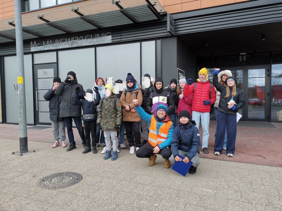 Grupa osób, w tym dzieci i dorośli, stoi razem przed budynkiem. Wszyscy są ciepło ubrani, co sugeruje chłodną pogodę. Dwie osoby kucają z przodu – jedna z nich nosi kamizelkę odblaskową. Panuje radosna atmosfera, grupa wygląda na gotową do wspólnej aktywności.