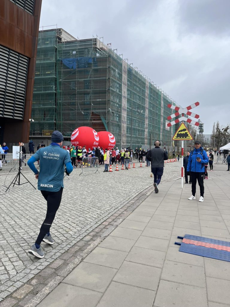 Scena z zawodów biegowych. Jeden z uczestników, z imieniem Marcin na plecach, biegnie w kierunku tłumu przy balonach Energa. W tle znajduje się budynek w remoncie otoczony rusztowaniem. Obok bieżni stoją osoby zabezpieczające trasę oraz obserwatorzy. Pogoda jest pochmurna.