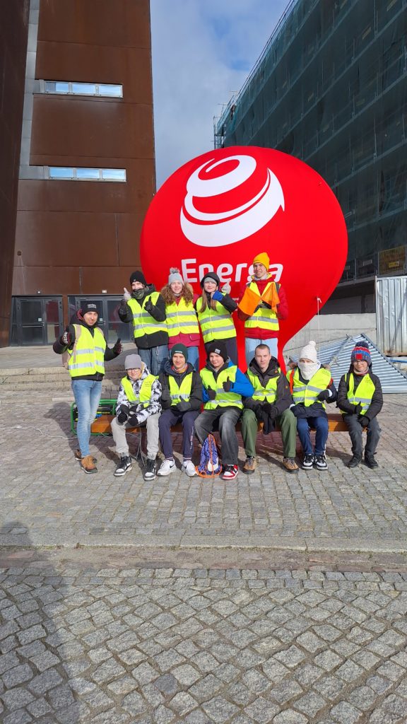 Grupa wolontariuszy w kamizelkach odblaskowych pozuje przed dużym balonem z logo Energa. Osoby w różnym wieku, większość uśmiechnięta i pokazująca kciuki w górę. Tło stanowi nowoczesny budynek oraz rusztowania. Atmosfera sugeruje udział w wydarzeniu sportowym.