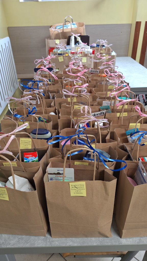 Na stole ustawiono równo wiele torebek papierowych z uchwytami, ozdobionych wstążkami. W każdej torbie znajdują się różne artykuły higieniczne i kosmetyczne, takie jak pasty do zębów, żele pod prysznic i dezodoranty. Do torebek przyklejono karteczki z życzeniami wielkanocnymi od uczniów z SP 47 w Gdańsku.