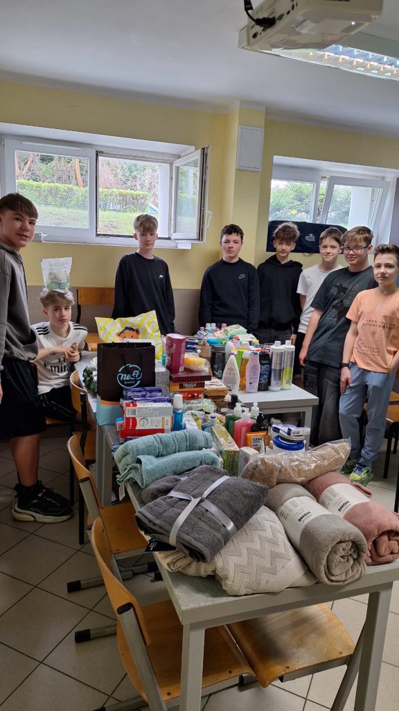 Grupa chłopców stoi wokół stołu w klasie, na którym zgromadzono liczne artykuły higieniczne, kosmetyki, ręczniki i środki czystości. Za nimi widać otwarte okna. Uczniowie najwyraźniej przygotowują się do pakowania darów.