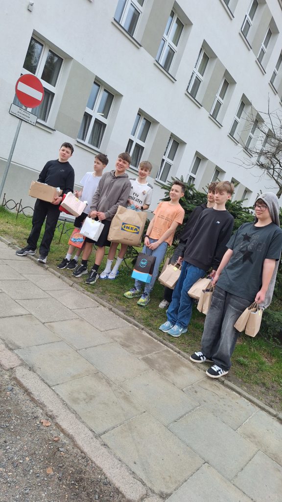 Kilku chłopców widać na zewnątrz budynku szkolnego, stojących przy chodniku z torbami w rękach. Uśmiechają się i trzymają różnorodne papierowe torby z upominkami, najwyraźniej gotowi do przekazania ich potrzebującym.