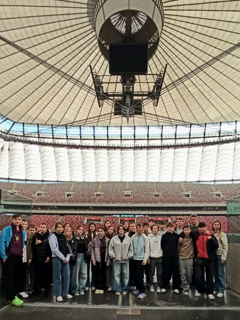 Uczniowie klasy 7b i 7c stoją na tle wnętrza Stadionu Narodowego. U góry jest promieniście roz-pięty dach z lin i membrany. Uczniowie stoją w centralnej części trybun, tuż przy krawędzi boi-ska, a za nimi widoczna jest ogromna widownia w kolorze czerwono-białym oraz centralnie za-wieszony ekran i konstrukcja oświetleniowa pod dachem stadionu.