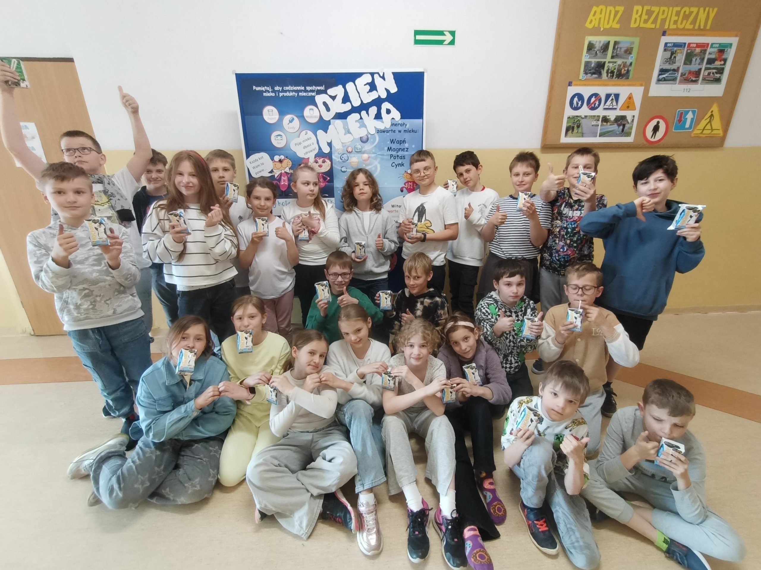 Dzieci z klasy 3a ustawione w dwóch rzędach na tle niebieskiej tablicy z hasłem "Dzień Mleka". Pierwszy rząd siedzi, a drugi stoi. Wszyscy trzymają w dłoniach kartonik z mlekiem.
