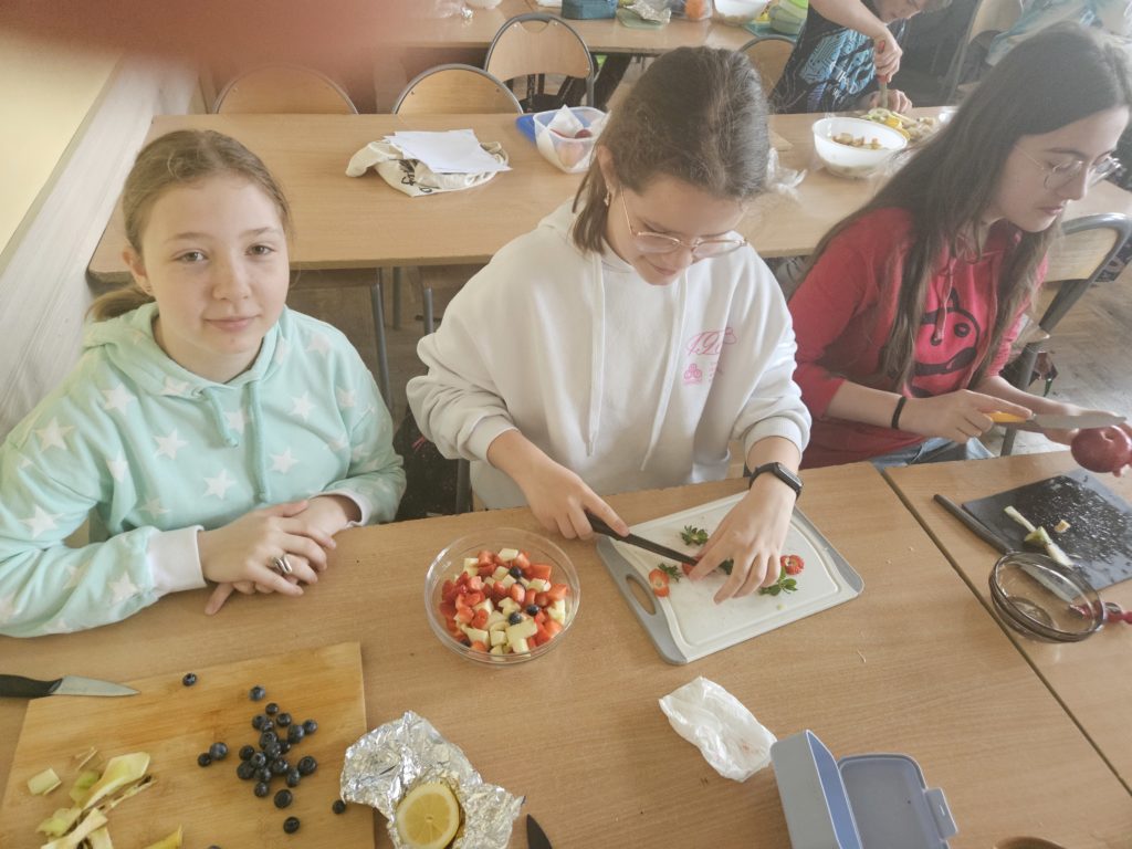 Dziewczynki na stołach przygotowują produkty do sałatki. Kroją umyte wcześniej owoce.