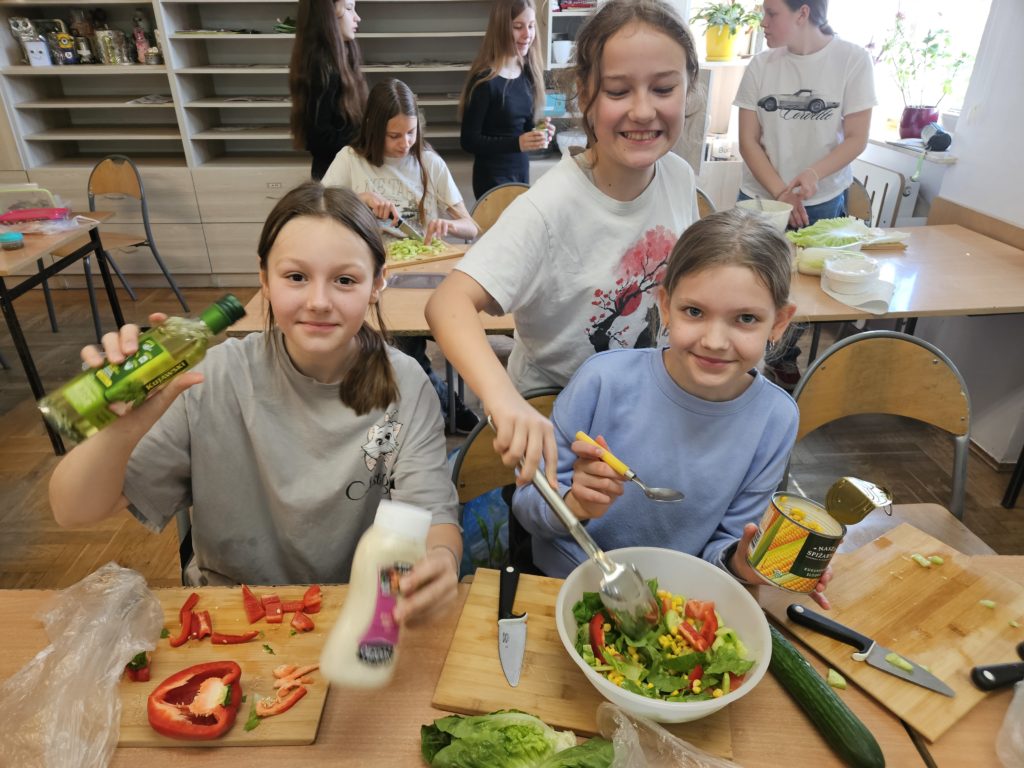 Dziewczynki na sole przygotowują warzywa do sałatki. Na stole stoją miseczki i deski z krojonymi warzywami.
