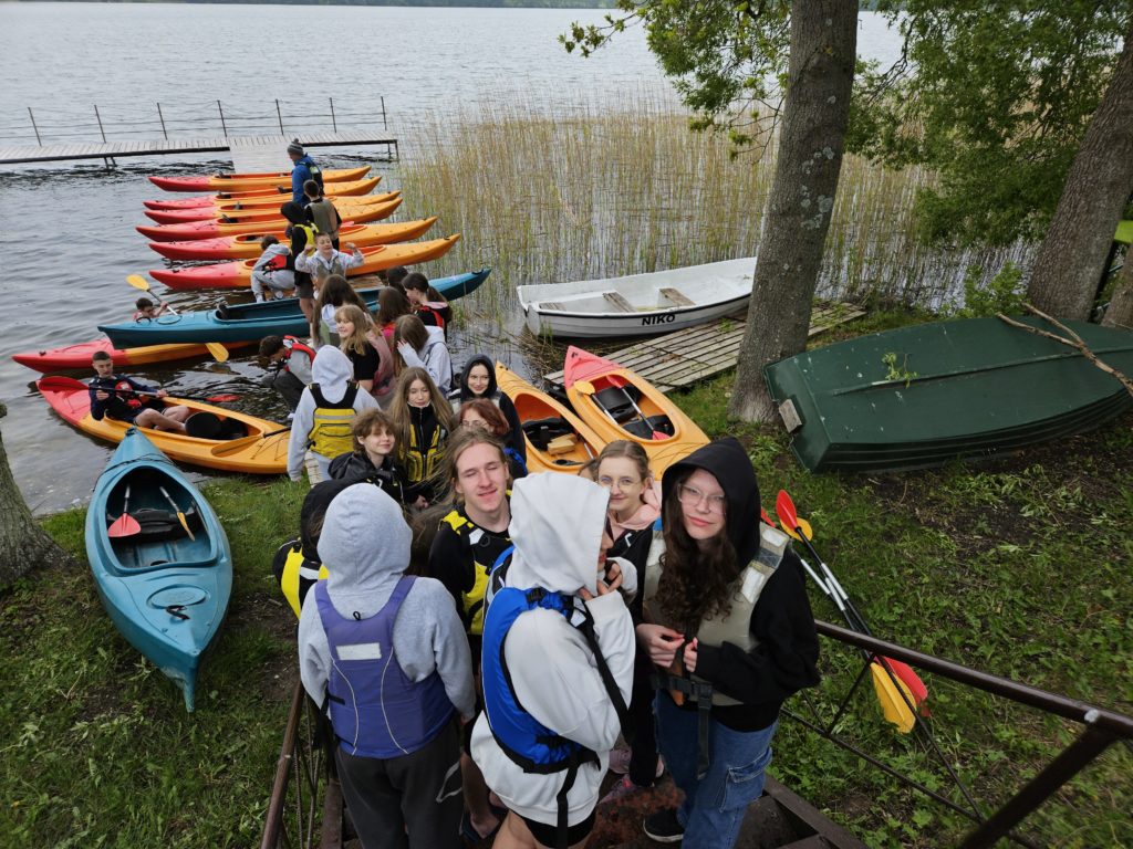 Na pierwszym planie młodzież schodząca po schodach do jeziora. Na jeziorze cumują kolorowe kajaki.