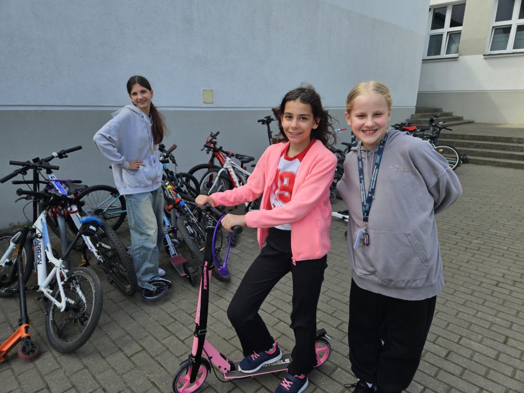 Dwie dziewczynki stoją przed szkołą. Jedna z nich na hulajnodze.