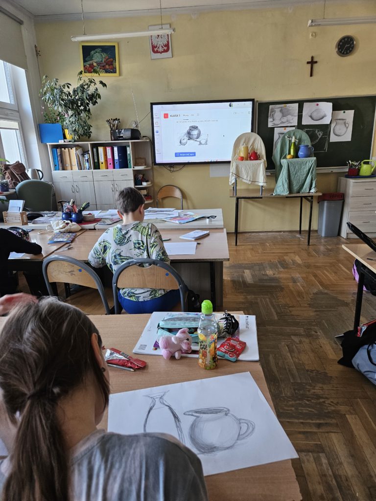 Na zdjęciu widać salę z ławkami przy których siedzą tyłem uczniowie rysujący ustawioną pod tablicą kompozycję różnych brył