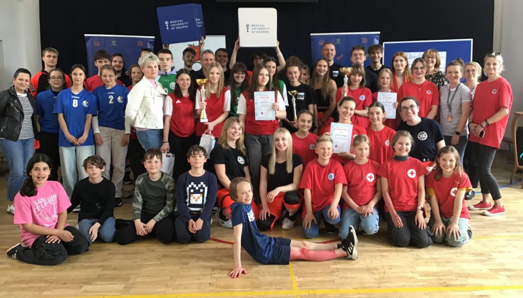 W sali gimnastycznej na tle banerów stoi ustawiona w kilka rzędów około pięćdziesięcioosobowa grupa ludzi: ratowników GUMed, uczniów, opiekunów drużyn i organizatorów konkursu z pierwszej pomocy. Trzy drużyny trzymają puchary i dyplomy.