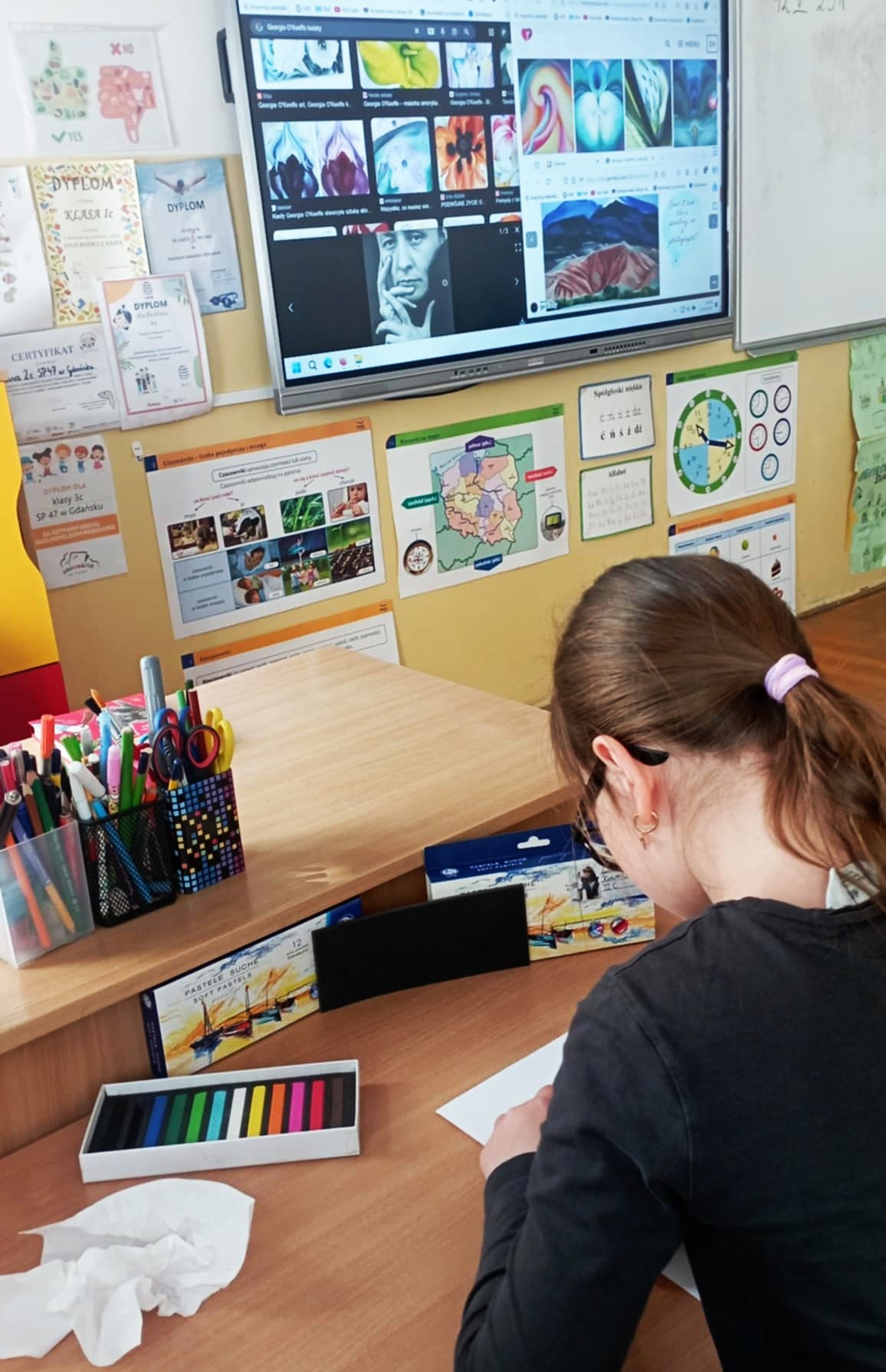 Na pierwszym planie dziewczynka rysująca na kartce suchymi pastelami, w tle tablica multimedialna wyświetlająca Georgię O'Keeffe i jej obrazy.