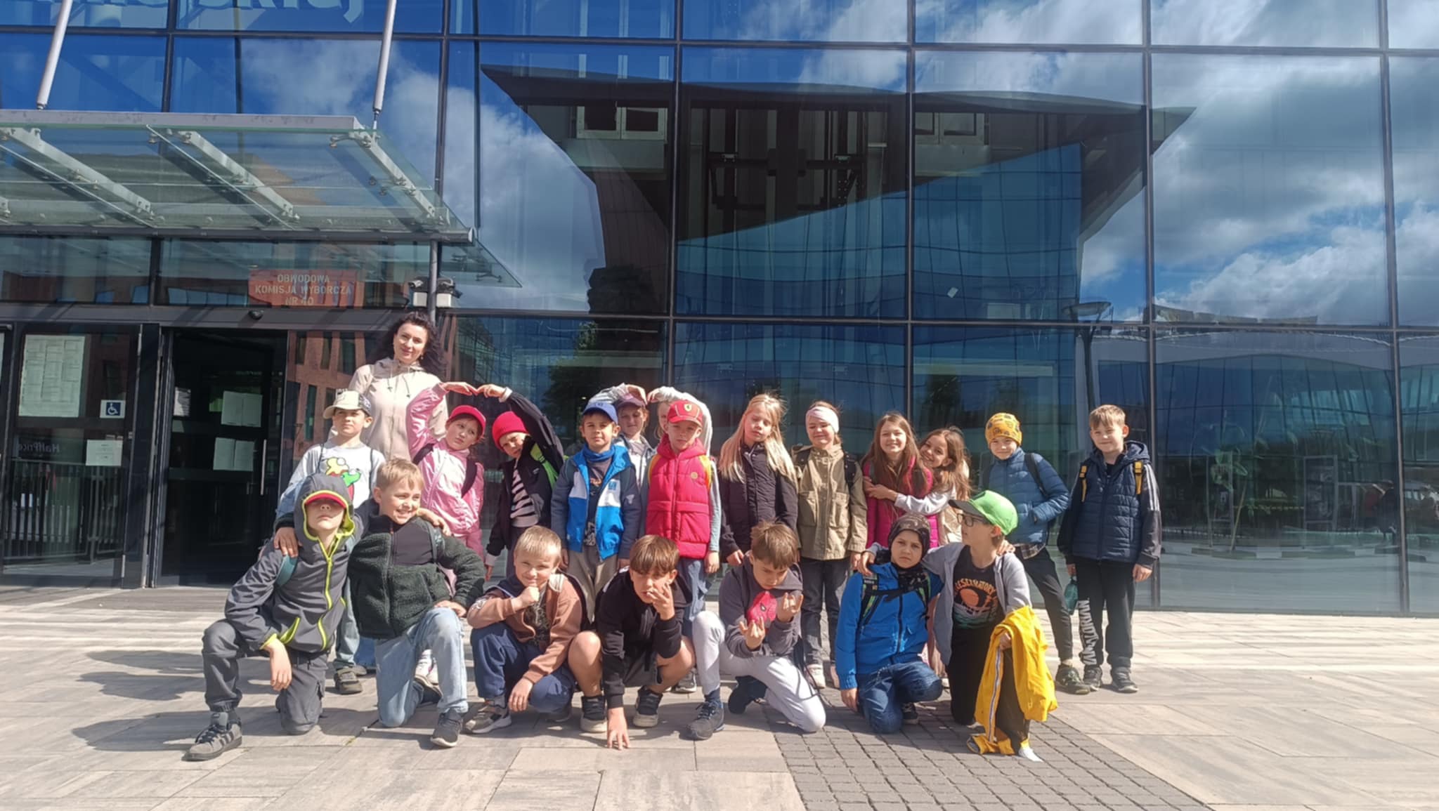 Grupa dzieci i dorosła kobieta stoją przed nowoczesnym budynkiem z przeszkloną elewacją. Dzieci są uśmiechnięte i ustawione w kilku rzędach. Kobieta stoi z tyłu, za dziećmi. W tle widać odbicie w szybach budynku.