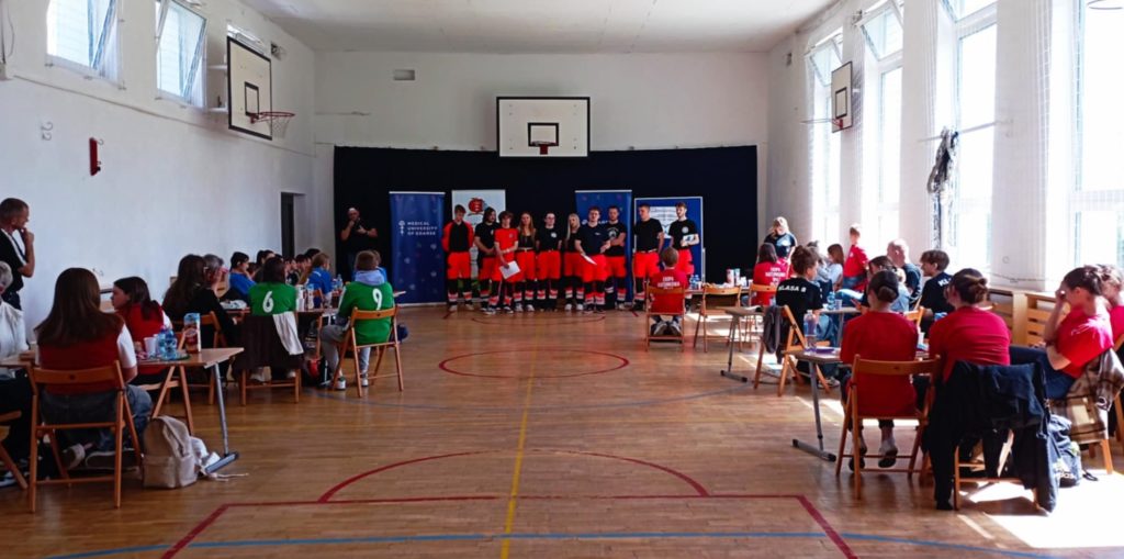 W sali gimnastycznej na tle banerów GUMed i SP 47 stoją ustawieni w rzędzie ratownicy medyczni sędziujący z zawodach. Przed nimi 6 trzyosobowych drużyn wraz z opiekunami siedzący w koło stolików.