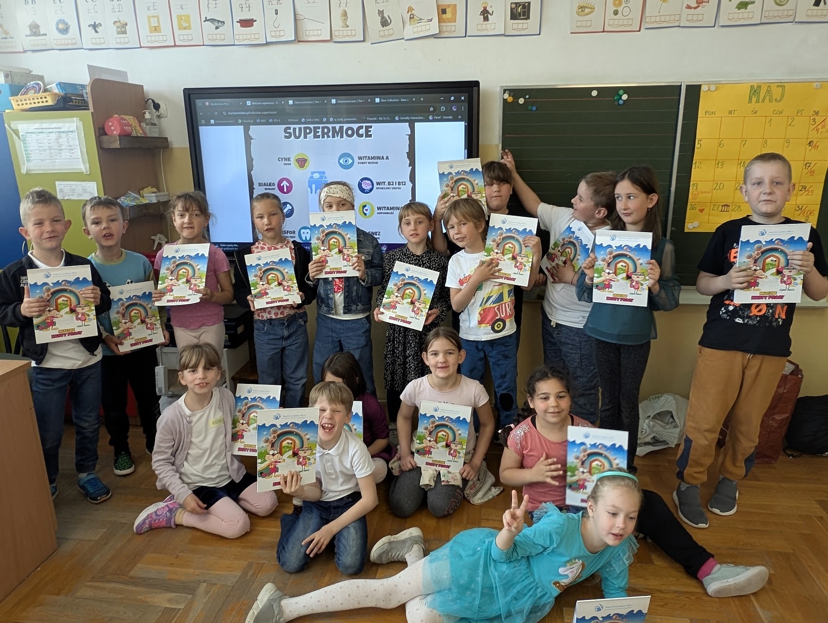 17 dzieci z klasy 1 c ustawionych na tle tablicy interaktywnej, gdzie wyświetla się napis "Supermoce" i informacje dotyczące wartości odżywczych mleka.  Wszyscy trzymają w dłoniach książeczki edukacyjne.