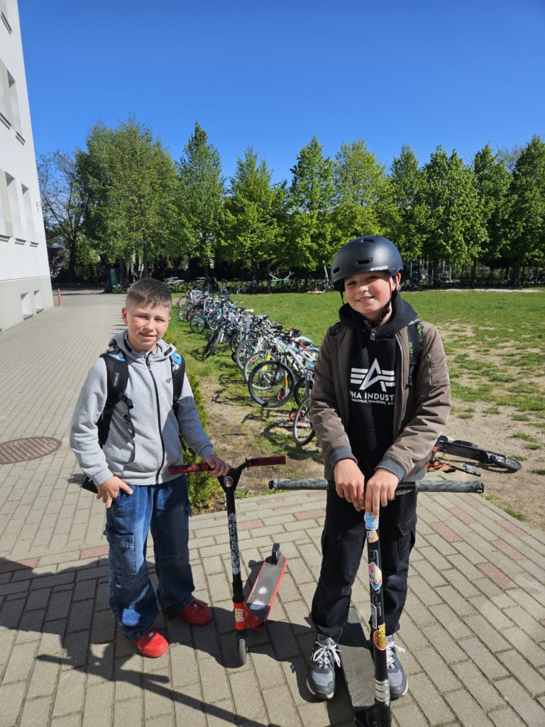 Dwaj chłopcy stoją z boku szkoły. jeden z nich trzyma hulajnogę. Za nimi stoją przypięte do stojaków rowery.