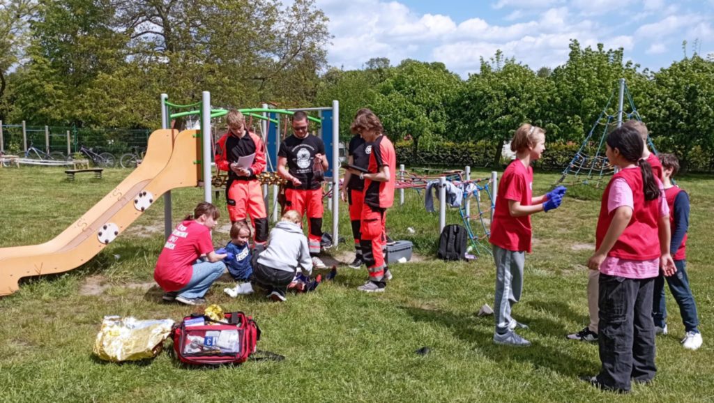 W szkolnym ogrodzie na tle zjeżdżalni stoi czterech sędziujących ratowników GUMed. Obok nich leży pozorujący chłopiec, któremu dwie uczennice udzielają pomocy, trzeci zawodnik z drużyny rozmawia ze świadkami zdarzenia.
