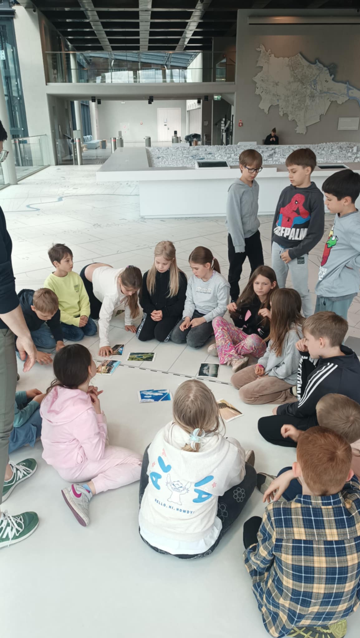 Grupa dzieci w wieku szkolnym siedzi w kręgu na podłodze w jasnym, nowoczesnym pomieszczeniu. Dzieci skupiają się na czymś w środku kręgu, prawdopodobnie na zdjęciach. W tle widać dorosłą osobę.