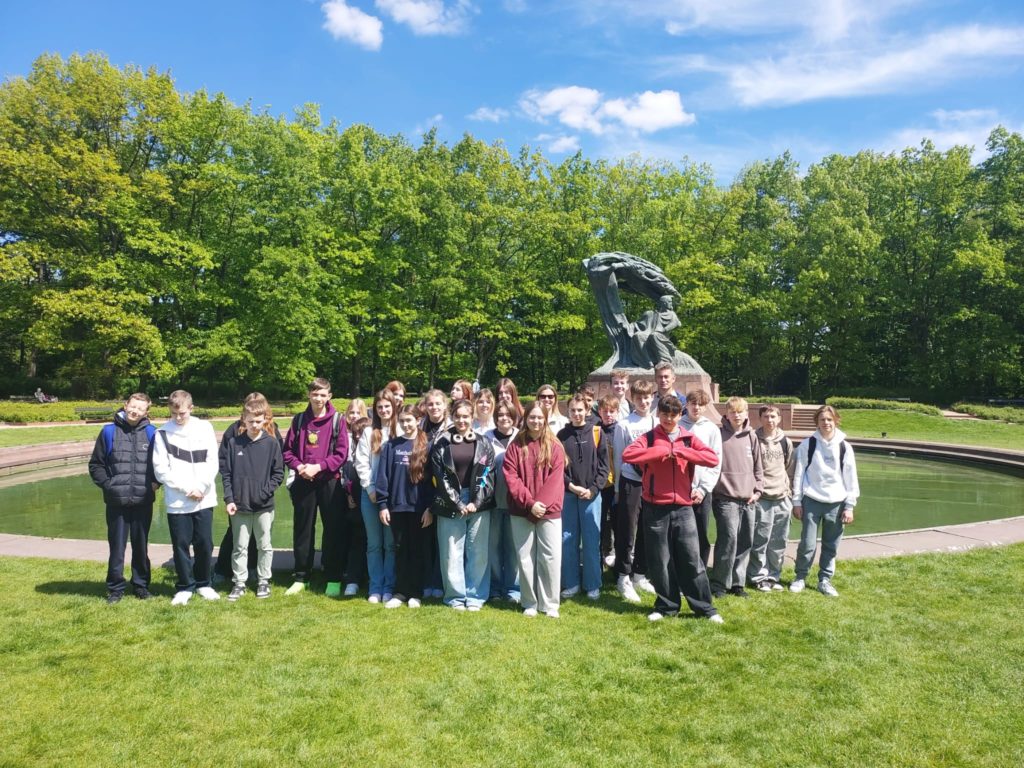 Uczniowie klasy 7b i 7b stoją na tle fontanny i pomnika Fryderyka Chopina w Łazienkach Kró-lewskich w Warszawie. Uczniowie stoją na zielonej trawie. W tle widać gęste, zielone drzewa oraz niebo z kilkoma chmurami.