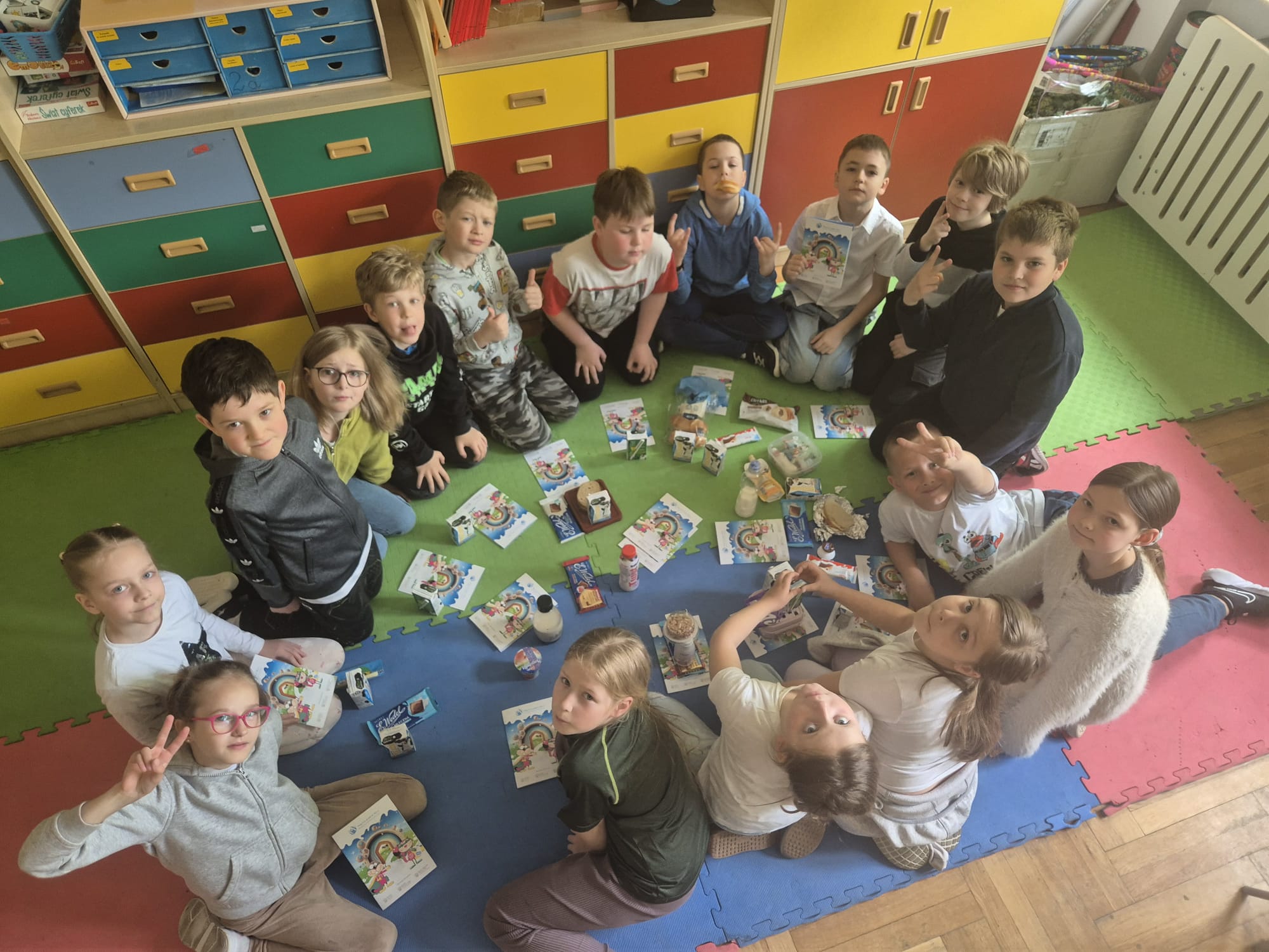 Dzieci siedzą na podłodze w kole. Przed nimi leżą książeczki edukacyjne o zaletach picia mleka oraz mleczne produkty np. jogurty, kefiry, serki i czekolady. W tle kolorowe szafki.
