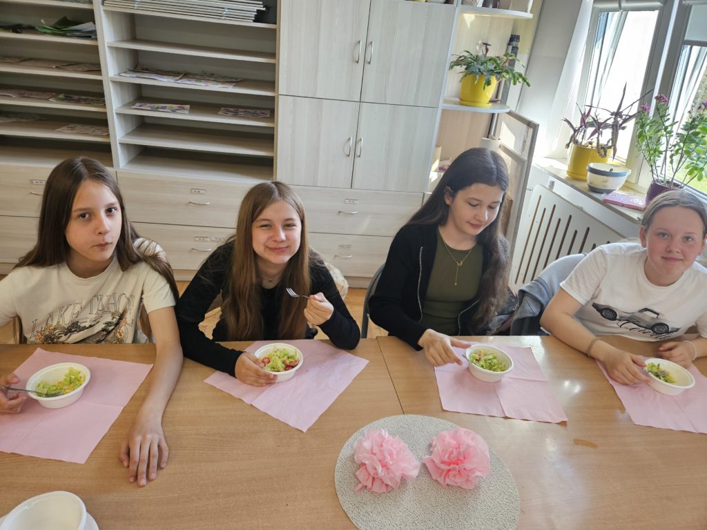 Dziewczynki siedzą za stołem, na którym znajdują się różowe dekoracje z serwetek oraz miseczki z nałożoną już sałatką.