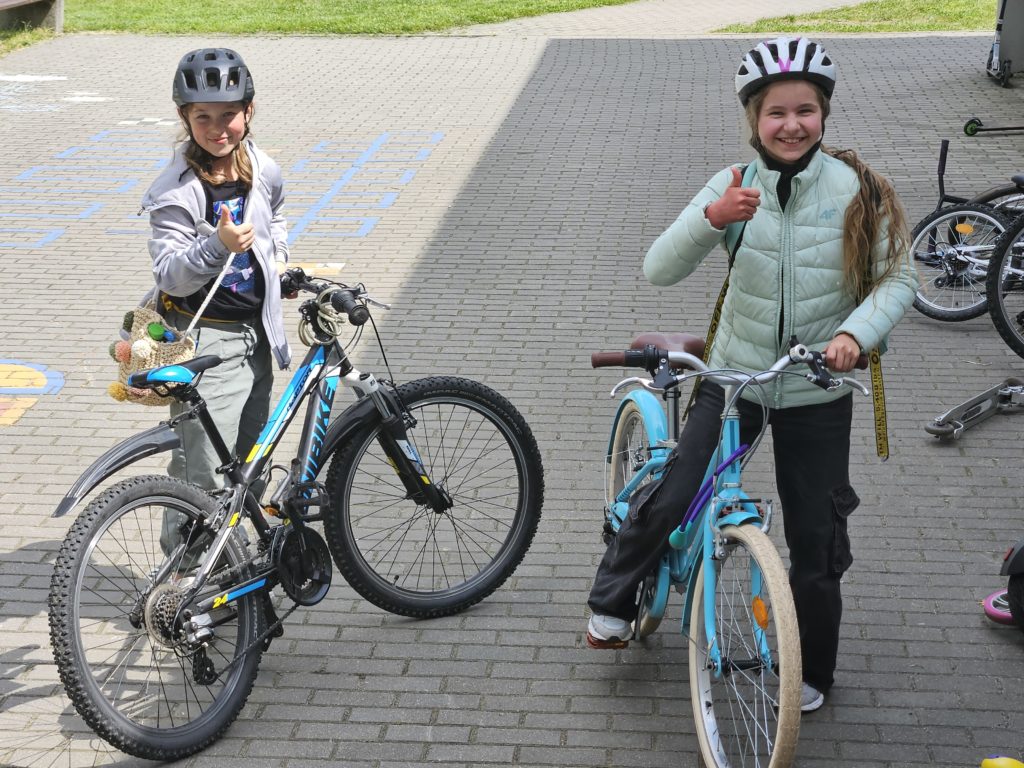 Dwie uśmiechnięte dziewczynki stoją z rowerami z boku szkoły. Obydwie trzymają swoje rowery. Na głowach maja założone kaski.