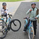 Dwie uśmiechnięte dziewczynki stoją z rowerami z boku szkoły. Obydwie trzymają swoje rowery. Na głowach maja założone kaski.