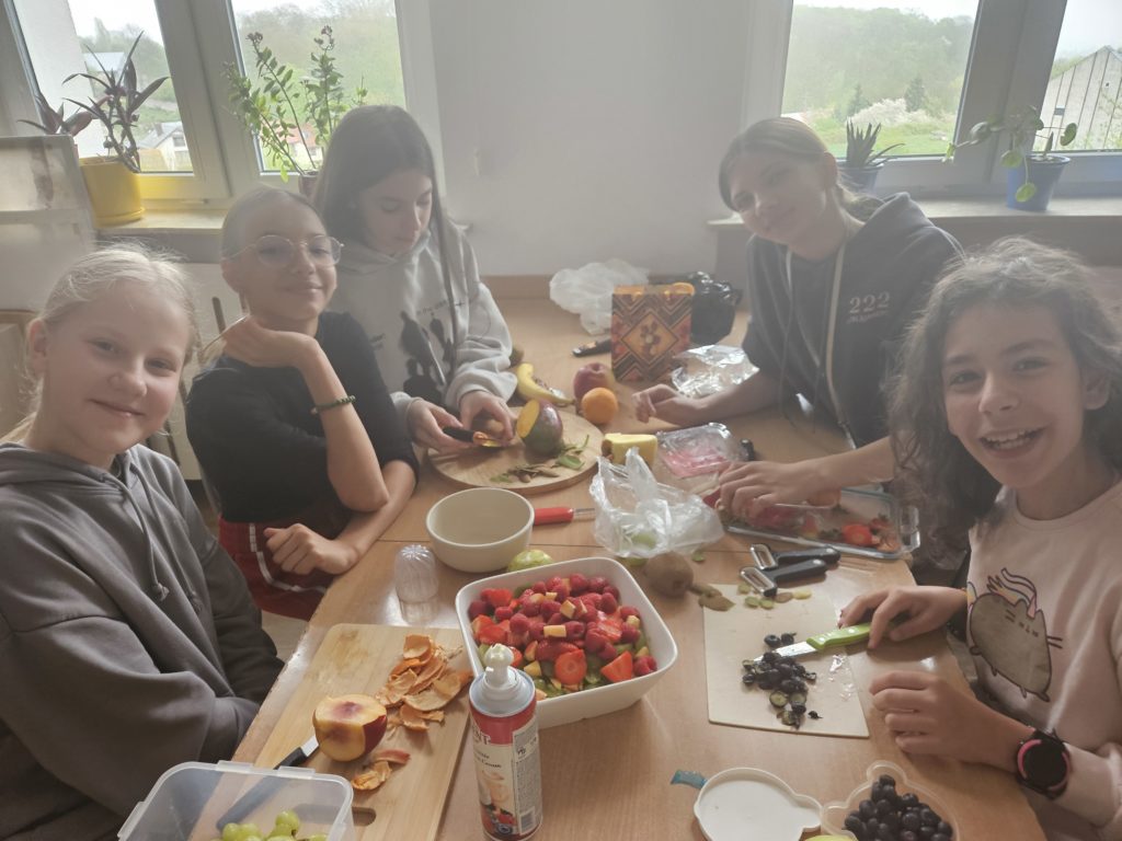 Na pierwszym planie stół a wokół niego dziewczynki przygotowujące owoce da sałatki.