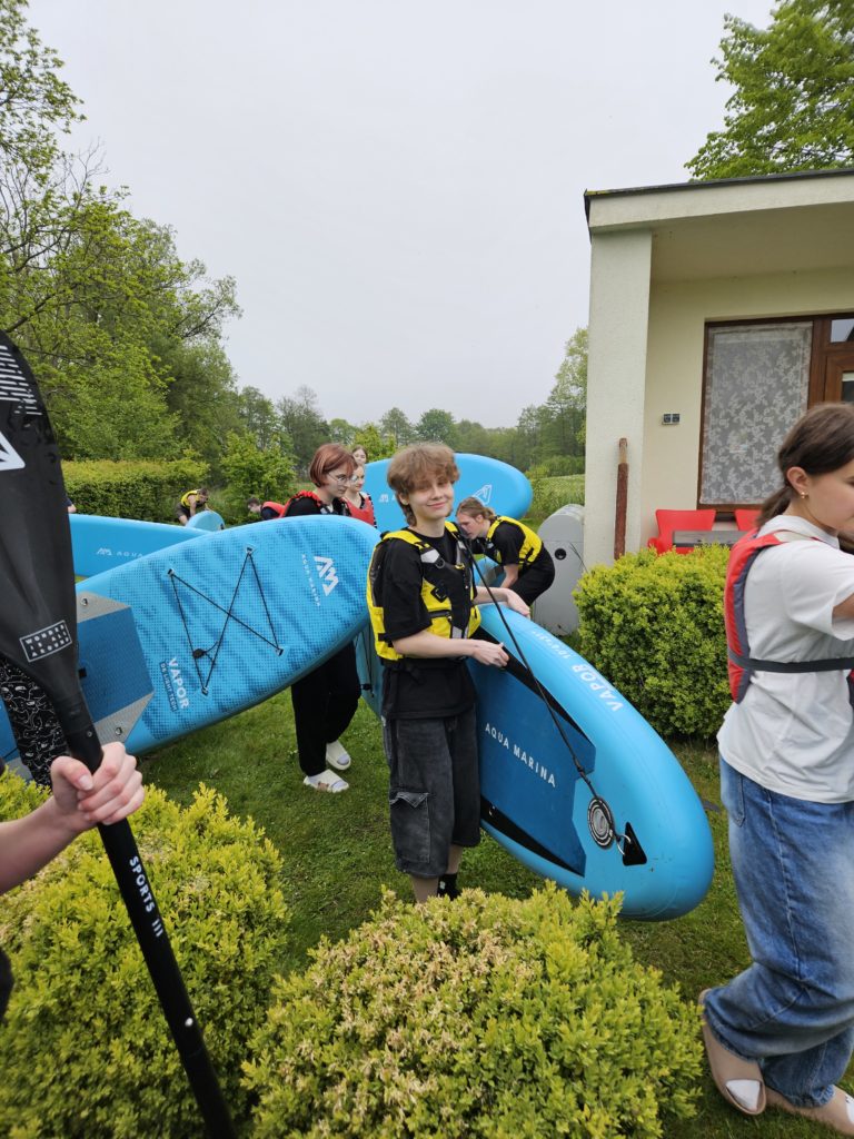 Ubrani w kamizelki pływacy wychodzą z niebieskimi deskami w kierunku jeziora