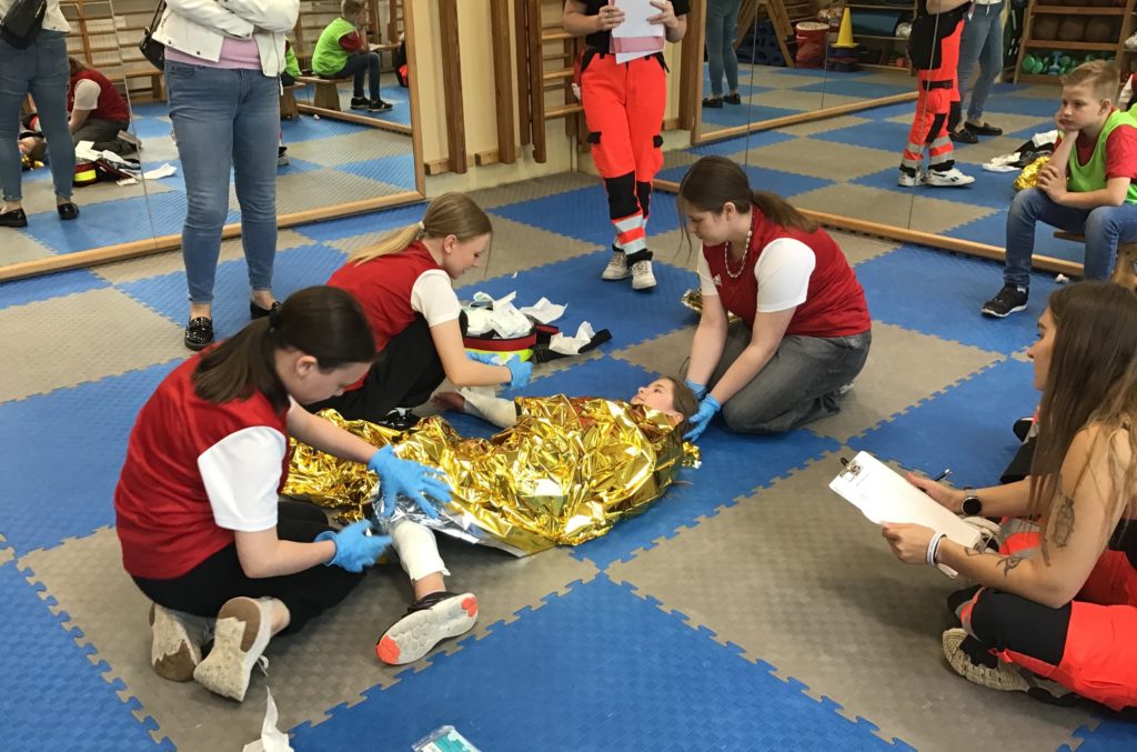 W sali gimnastyki korekcyjnej leży pozorująca dziewczyna, której trzy uczennice udzielają pomocy, dwie opatrują rany, a jedna stabilizuje głowę. W koło przyglądający się sędziowie zawodów i opiekun drużyny.