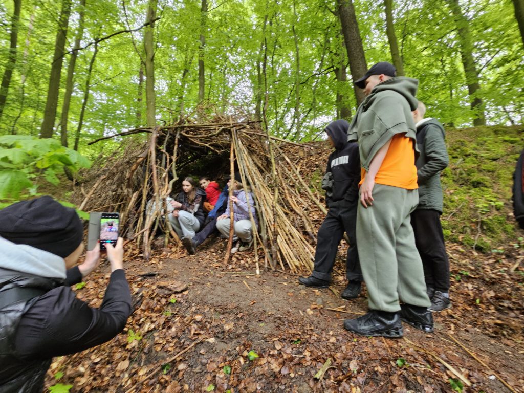 Na zdjęciu widać grupkę postaci przyglądającym się swojemu dziełu, czyli szałasowi zbudowanemu z kijów i liści
