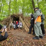 Na zdjęciu widać grupkę postaci przyglądającym się swojemu dziełu, czyli szałasowi zbudowanemu z kijów i liści