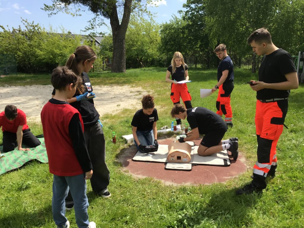 W szkolnym ogrodzie na pierwszym planie fantom, na którym uczniowie wykonują RKO z użyciem AED. Obok nich trzecia zawodniczka dzwoniąca po pomoc i rozmawiająca ze świadkiem zdarzenia. W koło ratownicy sędziujący w konkursie.