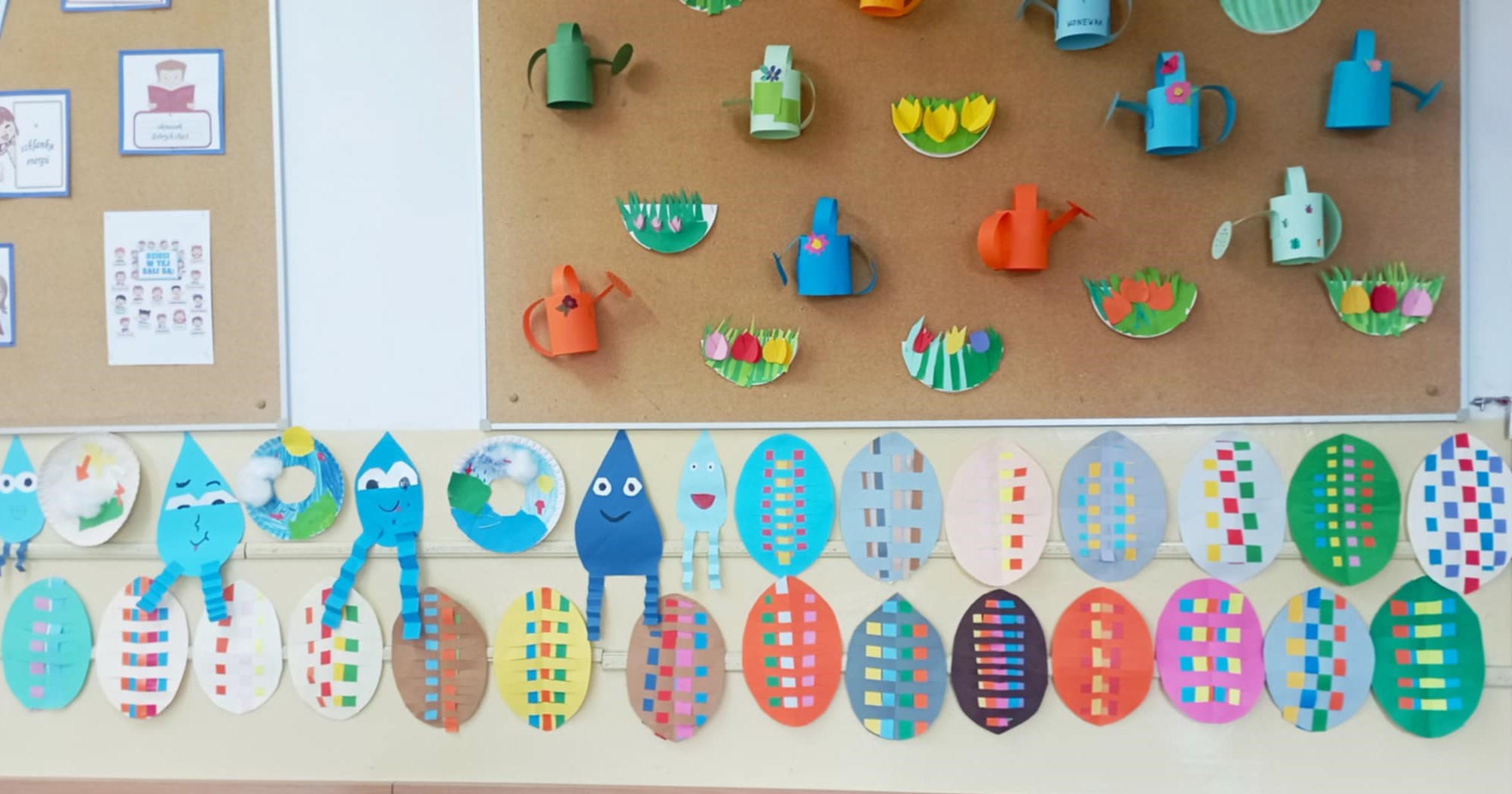 Na ścianie dwa rzędy freblowskich przeplatanek w kształcie jajek. Nad nimi tablica korkowa z wiosennymi kwiatami i konewkami w formacie 3d.