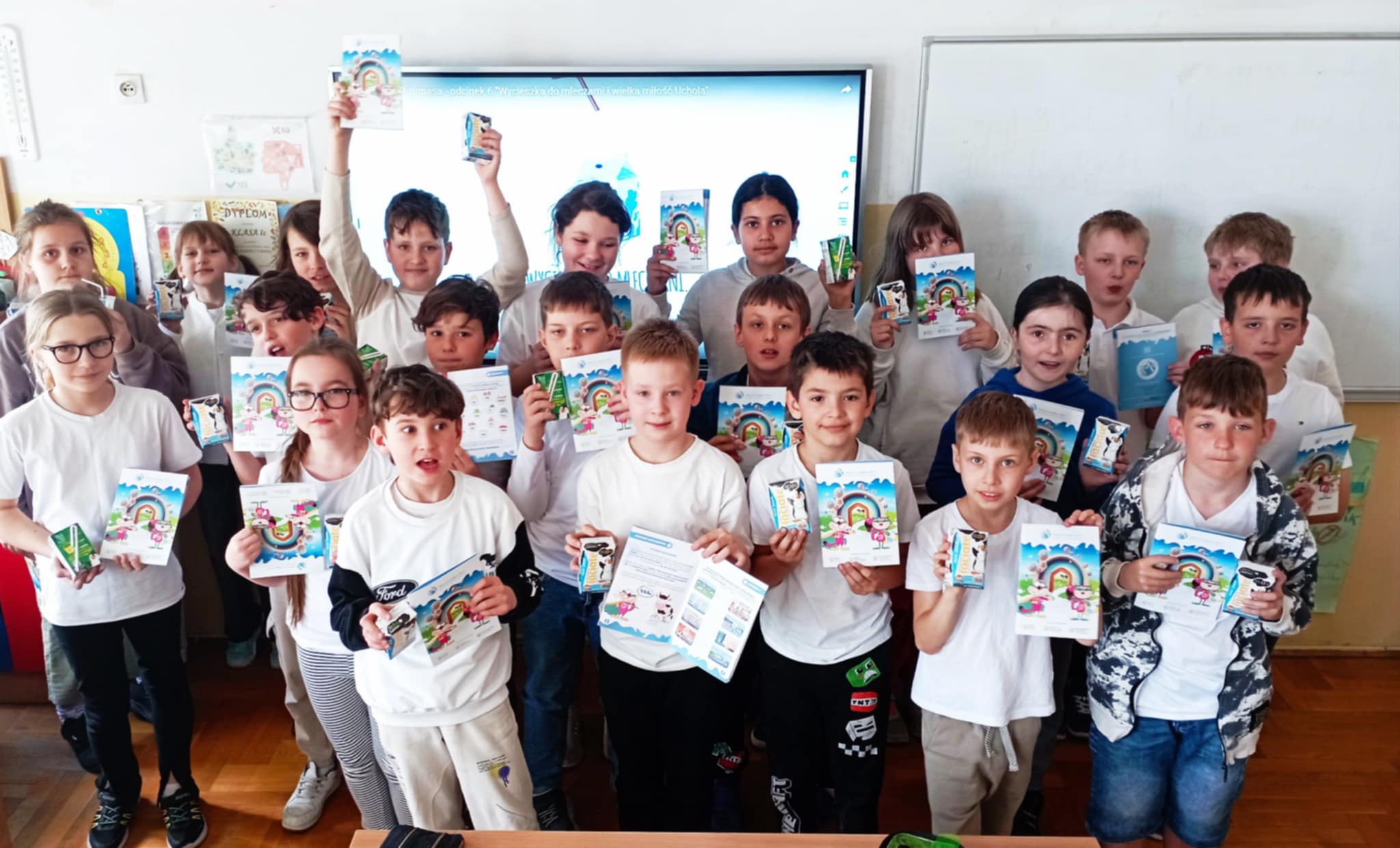 Uczniowie z 3c ustawieni w sali lekcyjnej na tle dwóch tablic. Wszyscy mają ubrane białe koszulki, a w dłoniach trzymają mleko i książeczki edukacyjne o mleku.