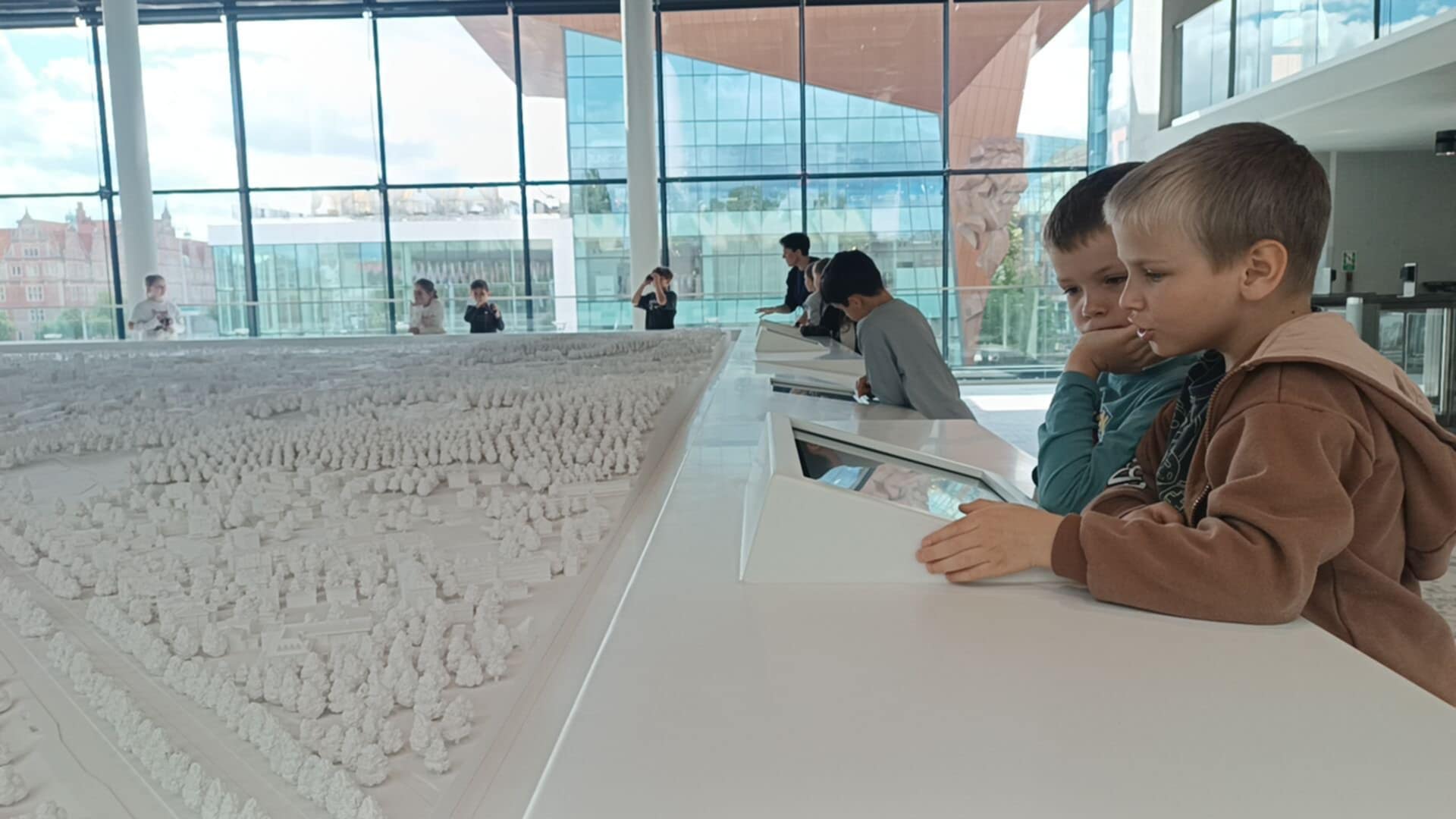 Dwóch chłopców ogląda dużą, szczegółową makietę miasta w nowoczesnym, przeszklonym wnętrzu muzeum lub centrum edukacyjnego. W tle widać inne dzieci oraz dorosłych obserwujących ekspozycję.