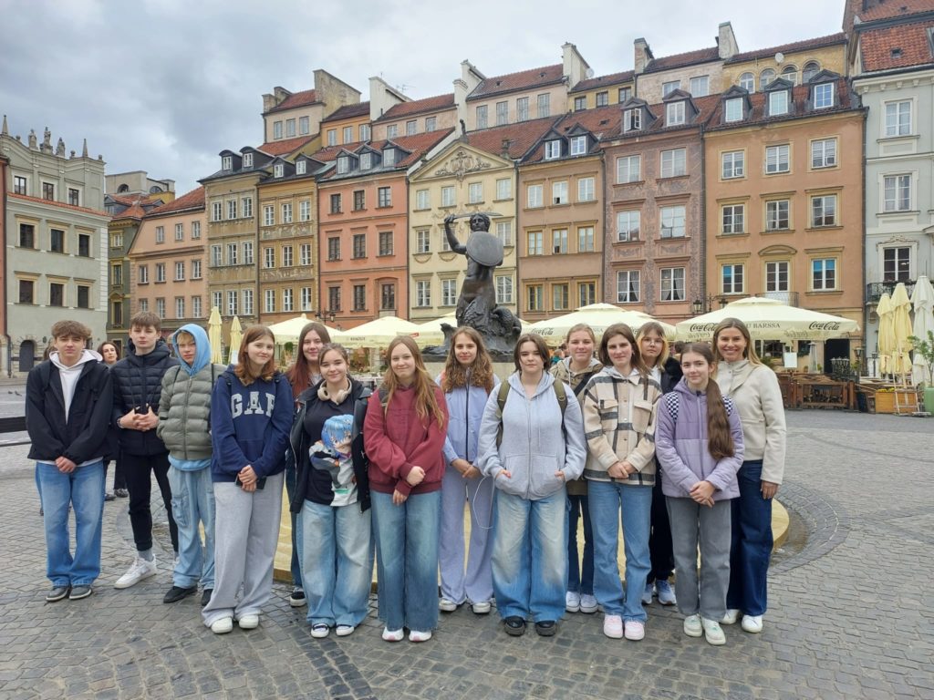 Uczniowie klasy 7c wraz z wychowawcą stoją na tle kolorowych kamienic w Starym Mieście w Warszawie. W centrum kadru znajduje się Syrenka Warszawska. W tle widać parasole kawiarni oraz brukowany plac