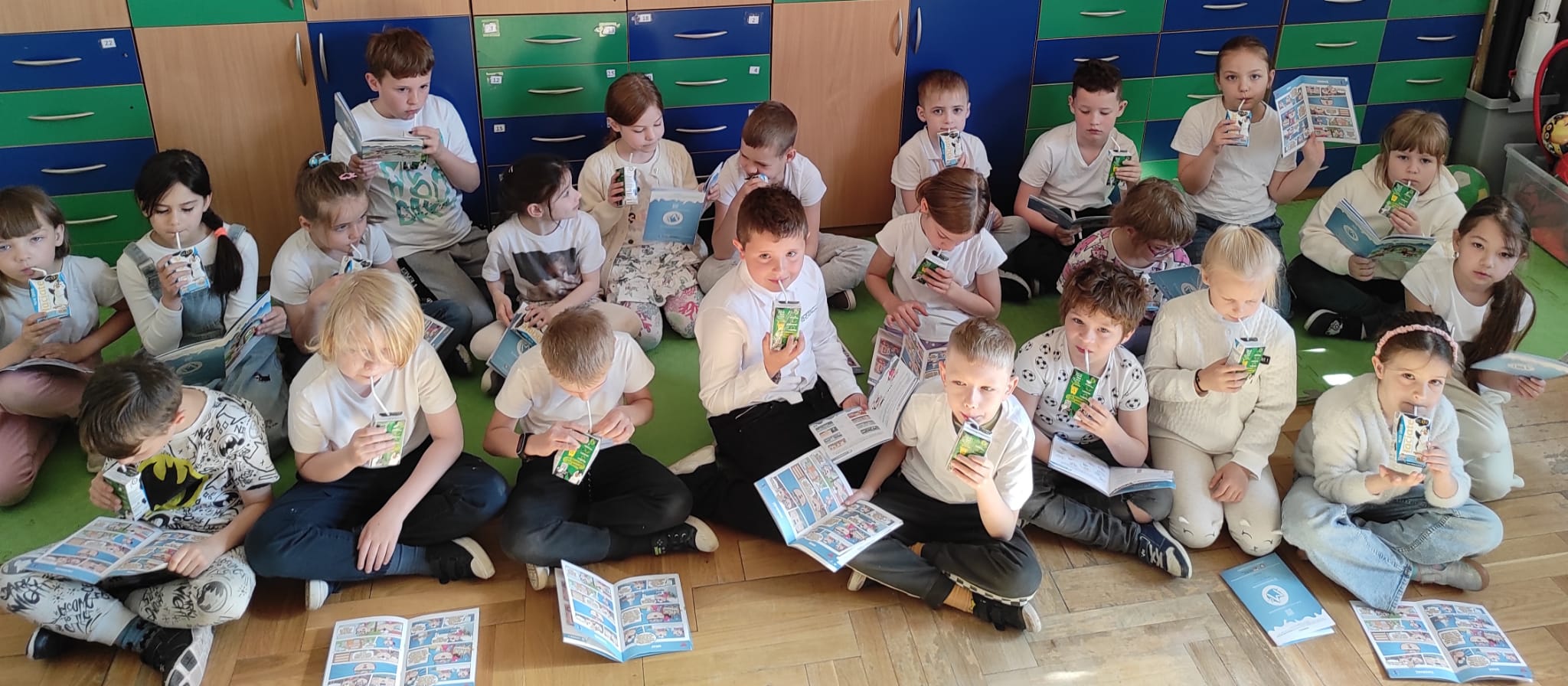 Uczniowie 1a siedzą na podłodze i czytają komiks o mleku. Wszyscy mają biały akcent w ubiorze i piją mleko.