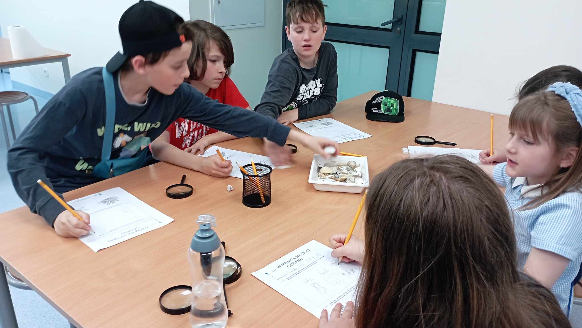Przy stole w laboratorium sześcioosobowa grupa uczniów klasy 3c notuje na karcie notatkę z obserwacji przyrodniczych. Na stole lupy i preparaty zwierząt.