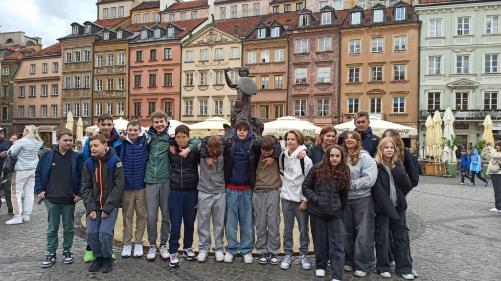 Uczniowie klasy 7b wraz z wychowawcą stoją na tle kolorowych kamienic w Starym Mieście w Warszawie. W centrum kadru znajduje się Syrenka Warszawska. W tle widać parasole kawiarni oraz brukowany plac.