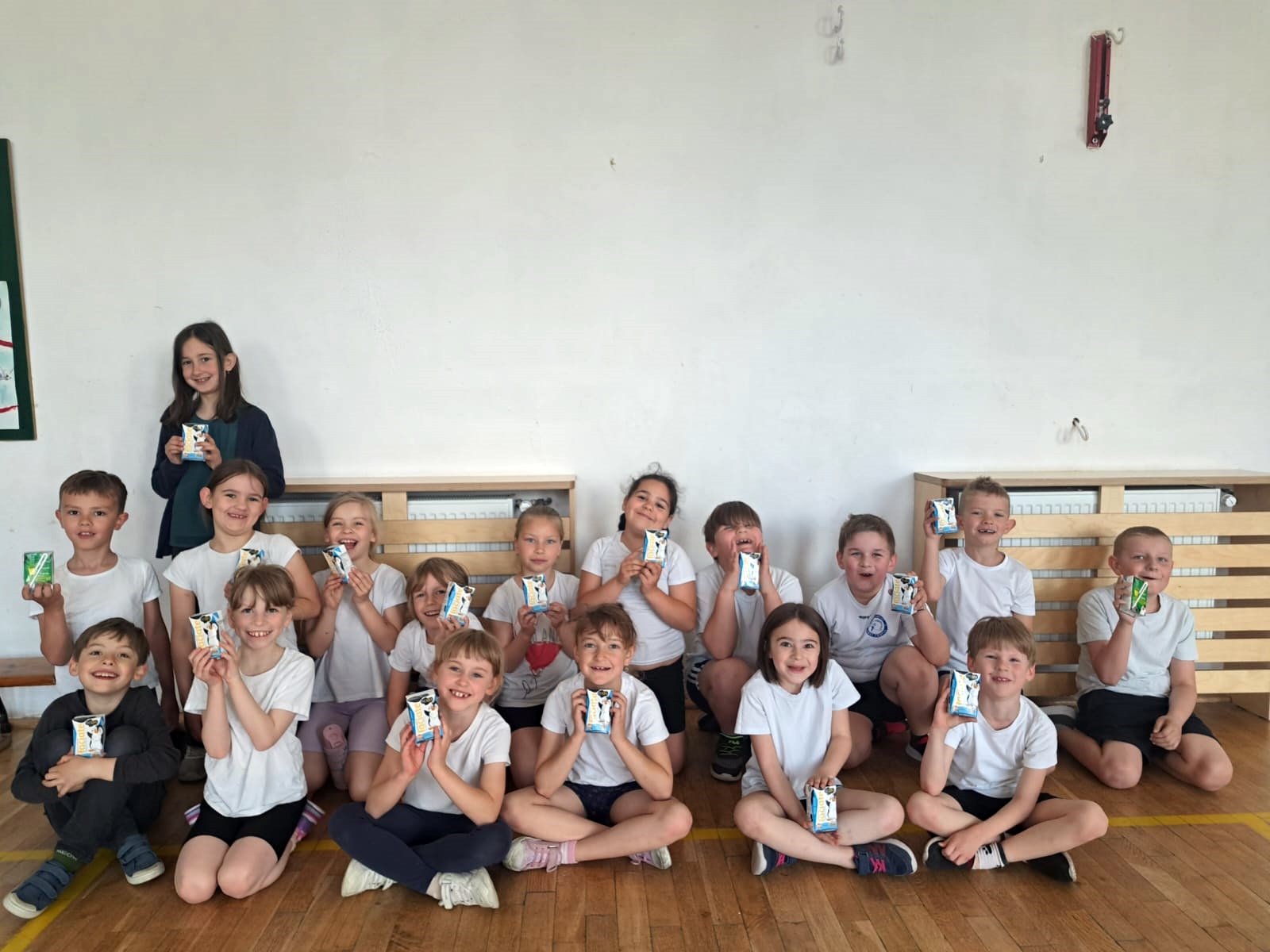 16 uczniów z 1c siedzi na podłodze w sali gimnastycznej. Jedna dziewczynka w ostatnim rzędzie stoi. Wszyscy są uśmiechnięci i w dłoniach trzymają kartoniki z mlekiem.