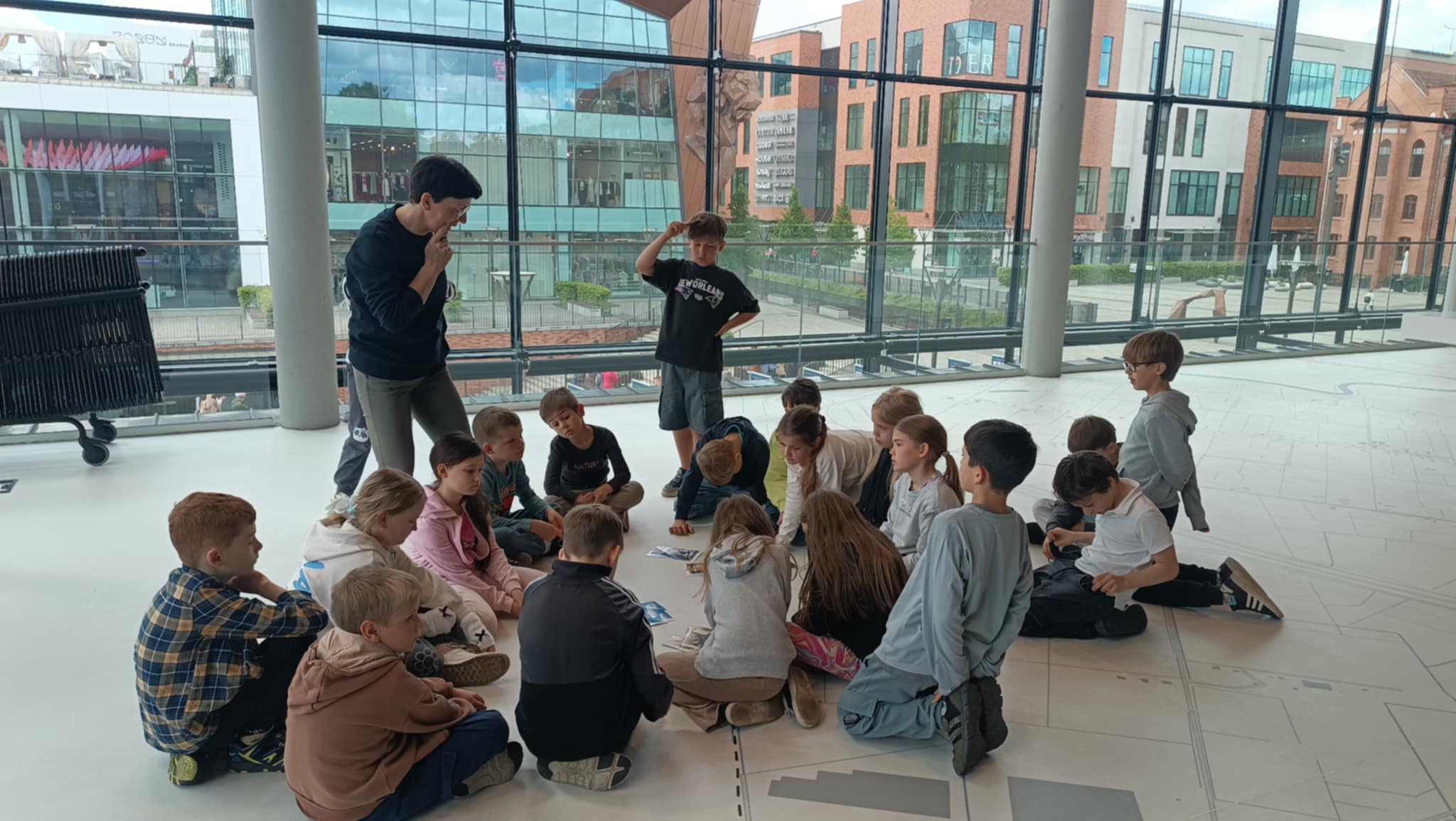 Dorosła kobieta i grupa uczniów siedzą w kręgu na podłodze w jasnym, nowoczesnym budynku z dużymi oknami, za którymi widać zabudowę miejską. Uczniowie skupiają się na czymś w środku kręgu. Atmosfera jest skupiona i spokojna.