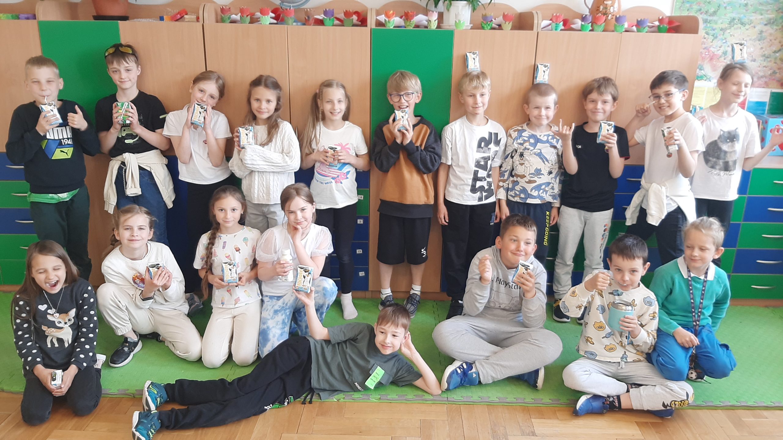 19 uczniów z 2a pozuje na tle szkolnych szafek. Dzieci są ustawione w dwóch rzędach. Pierwszy rząd siedzi, a drugi stoi. Wszyscy są uśmiechnieci i w dłoniach trzymają kartoniki z mlekiem.