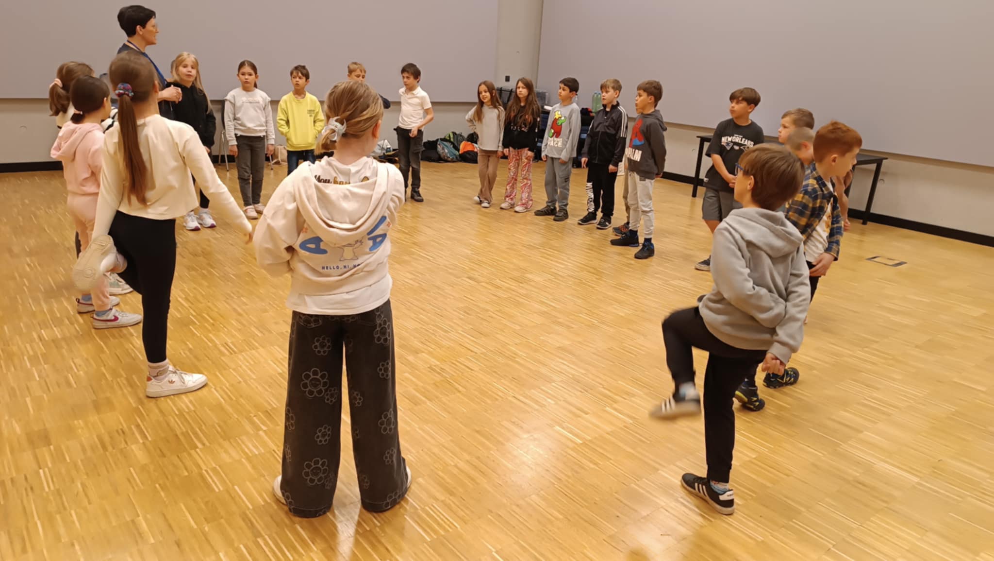 Grupa dzieci stoi w kręgu na parkiecie w sali gimnastycznej. W kręgu znajduje się również również dorosła kobieta. Dzieci wydają się wykonywać ćwiczenia lub rozgrzewkę. Podłoga jest drewniana.